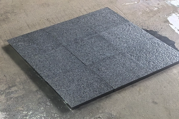 Dark-grey G654 Granite paving panel shown during visual surface review on a wet concrete floor